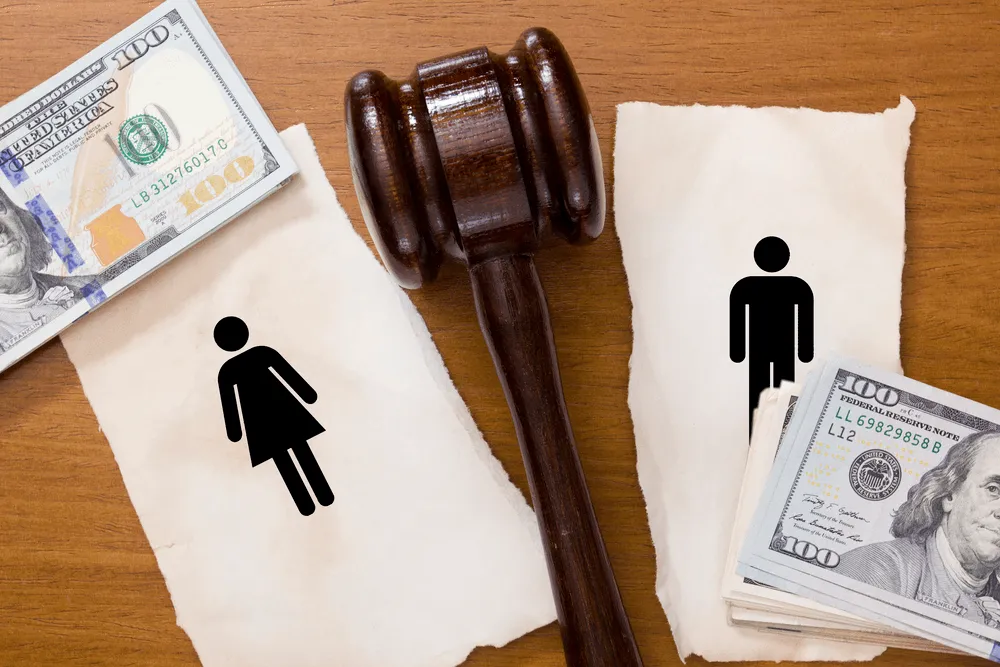 A gavel in the middle of a ripped piece of paper that has the image of woman on one side and a man on the other. A stack of money is on either side. It is representing financials in a divorce.