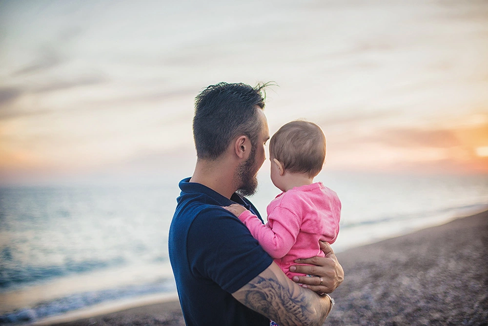 Father holding his child on the beach at sunset. Protect your rights as a father in Sugar Land with help from our paternity rights divorce lawyer.