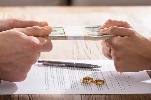 Divorce paperwork with a pen and wedding rings on top with two sets of hands pulling at a stack of bills representing fighting over money. 