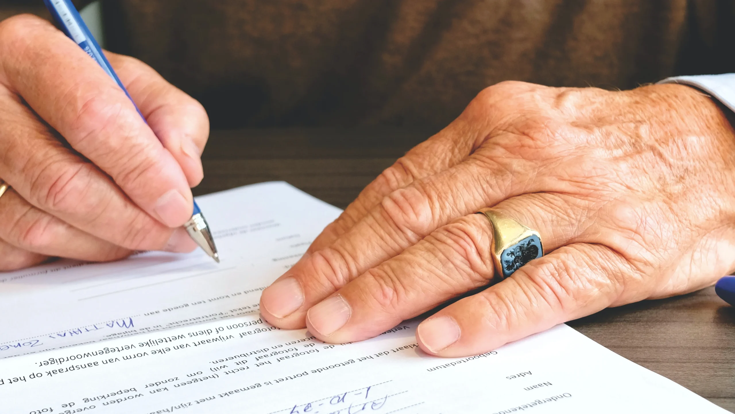 Close up of an older man's hands as they sign some papers. He has on a gold ring. 